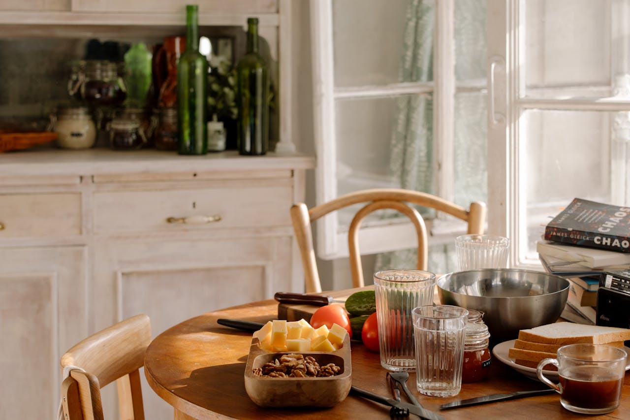 A sunlit kitchen table with food, glassware, and books, creating a warm, inviting ambiance.